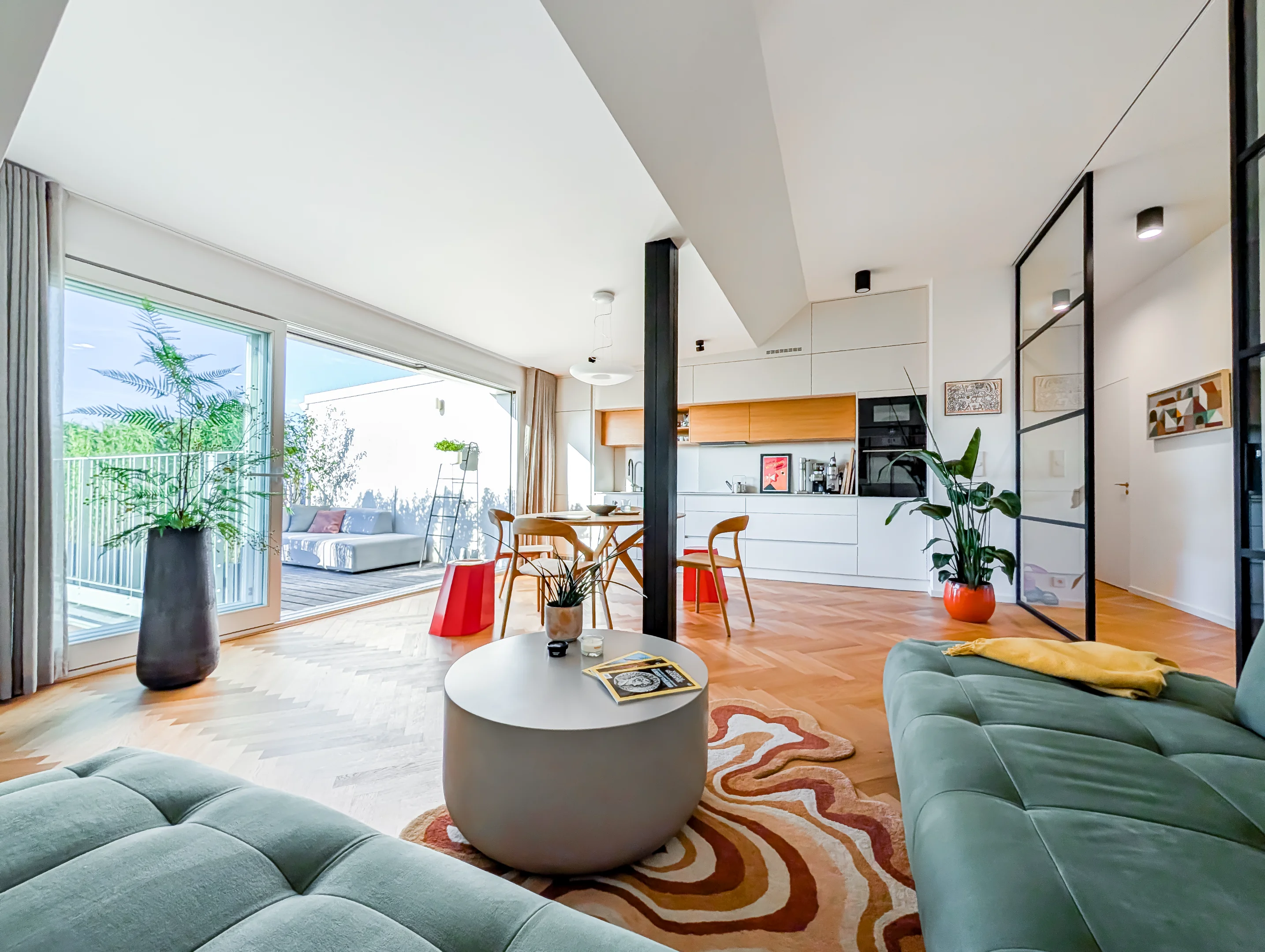 Světlý interiér: obývák s kuchyní, sedačka, dřevěná podlaha a zeleň.